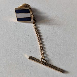 May include: A gold-coloured tie pin with a square enamel design. It has a blue top and bottom border with a white stripe in the middle. A fine gold chain connects the square to a horizontal bar.
