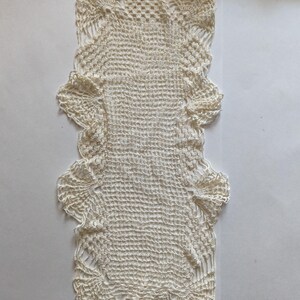 May include: Ivory-colored, rectangular crochet doily with a textured, open weave design. The doily features ruffled edges and a symmetrical pattern. This handmade piece is suitable for home decor.