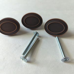 Puede incluir: Tres pomos de armario de forma ovalada con acabado en cobre y un borde de cuentas decorativas. Cada pomo viene con un tornillo plateado.