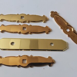 May include: Five gold-colored metal decorative hardware pieces. Two pieces are ornate with a square cutout in the center. One piece is rectangular with rounded corners and two circular cutouts. Two pieces are ornate with a square cutout in the center.