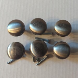 May include: A set of six round, brushed nickel cabinet knobs with a smooth, metallic finish. Each knob features a circular face and a central mounting point on the back. Several silver-coloured metal screws are included for installation.