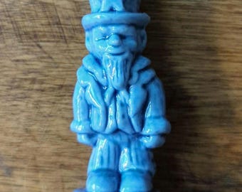 May include: A small, light blue ceramic figurine depicting a figure wearing a tall top hat with a prominent star on the front, a long coat, and striped trousers. The character has a pointed beard and is standing on a circular base.