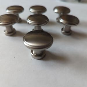 Könnte beinhalten: Eine Sammlung von sieben Schrankknöpfen aus gebürstetem Nickel. Jeder Knopf hat eine runde, flache Oberseite und einen kurzen, zylindrischen Sockel. Die Knöpfe sind auf einer weißen Oberfläche angeordnet und zeigen ihr schlichtes, modernes Design. Geeignet für Schubladen und Schränke.