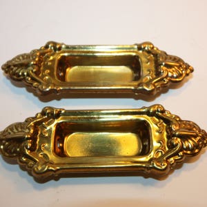 Könnte beinhalten: Zwei verzierte goldfarbene Metall-Tür- oder Fenstergriffe mit einem rechteckigen Ausschnitt in der Mitte. Die Griffe haben ein dekoratives, verziertes Design mit einem blattartigen Muster.