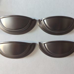Puede incluir: Cuatro tiradores de armario de color bronce dispuestos en forma cuadrada. Cada tirador tiene un diseño semicircular con una parte superior curvada y un centro ligeramente hundido. Los tiradores son de metal y tienen un acabado liso y mate, adecuados para armarios de cocina o baño.