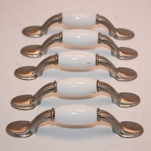 May include: Five white ceramic cabinet knobs with a silver finish. The knobs are oval shaped and have a slight curve.