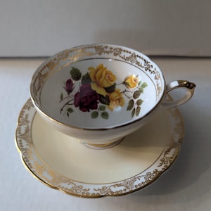 May include: A vintage teacup and saucer set. The teacup has a floral design with yellow and burgundy roses. The rim and handle are trimmed with gold. The saucer is a pale yellow with a gold filigree border.