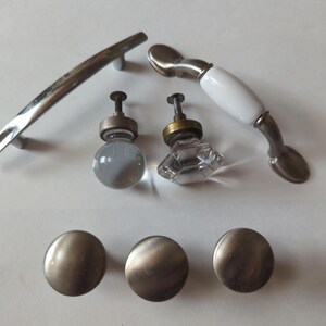 May include: A collection of cabinet hardware, including a long, curved silver handle, a white and silver handle, two glass knobs, and three round, brushed nickel knobs. These pieces offer a variety of styles for furniture upgrades.