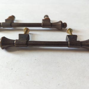 Puede incluir: Dos tiradores de armario antiguos de color bronce con extremos decorativos y una barra central. Cada tirador tiene un tornillo de latón para el montaje. Ideales para la restauración de muebles o para dar encanto vintage.