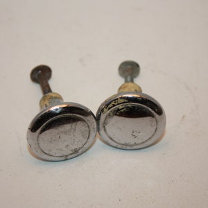 May include: Two silver-toned cabinet knobs with a round, slightly raised, center and a slightly raised outer edge. The knobs have a screw-on back.