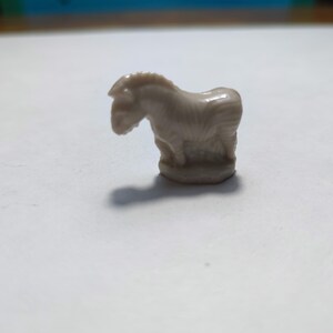 May include: A small, white ceramic figurine of a zebra standing on a base. The zebra has a striped pattern and is facing to the left.