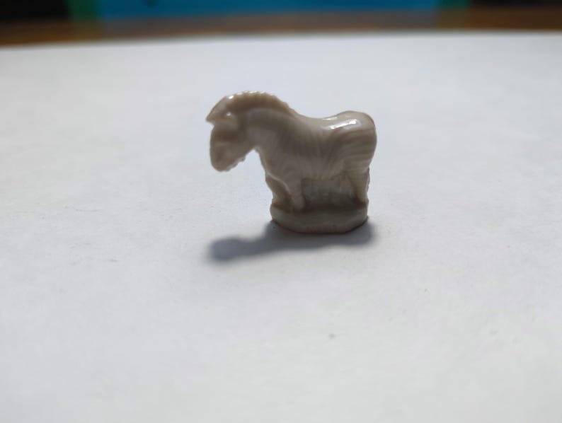 May include: A small, white ceramic figurine of a zebra standing on a base. The zebra has a striped pattern and is facing to the left.