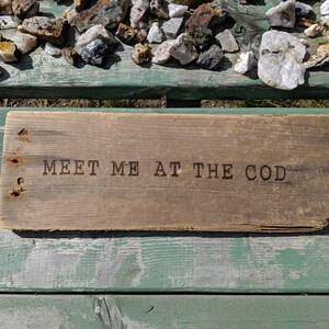 Op de afbeelding: Een rustiek houten bord met de tekst "MEET ME AT THE COD" gebrand in het oppervlak. Het bord ligt op een verweerde groene houten bank.