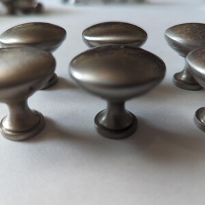 Op de afbeelding: Een verzameling van zes ovale kastknoppen met een geborstelde nikkel afwerking. De knoppen hebben een klassiek ontwerp, met een afgeronde bovenkant en een voet. Geschikt voor laden en kasten.