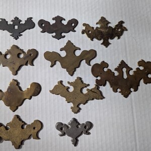 May include: A collection of 11 ornate brass drawer pulls with intricate designs. The pulls are in various shapes and sizes, with some featuring keyholes.