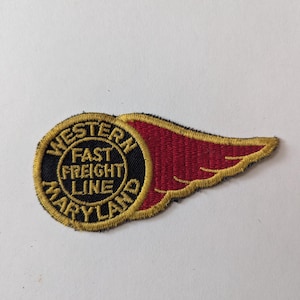 May include: Embroidered patch featuring the text "WESTERN FAST FREIGHT LINE MARYLAND" in gold lettering on a black circle, with a red wing design. The patch is likely for a vintage train or transportation company.