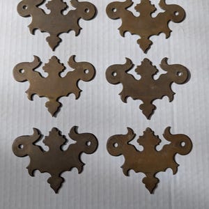 Op de afbeelding: Zes antieke messing decoratieve hardware stukken met ingewikkelde bloemmotieven. Elk stuk heeft een unieke vorm en beschikt over een centraal bloemmotief omgeven door zwierige, sierlijke details.