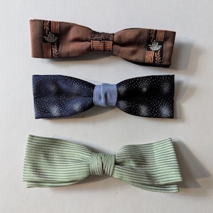 May include: Three bow ties in a row. The top bow tie is brown with a striped pattern and a small emblem. The middle bow tie is navy blue with white dots and a light blue center. The bottom bow tie is light green with thin stripes.