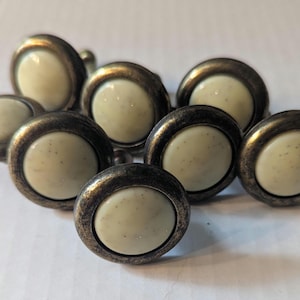 May include: A collection of seven round cabinet knobs featuring a smooth, off-white speckled center inlay surrounded by a dark, distressed metallic rim with a weathered finish. Each knob has a circular profile and a raised, domed surface design.