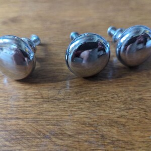 May include: Three shiny, chrome-finished cabinet knobs with a rounded, oval shape. The knobs are arranged on a wooden surface, reflecting the surrounding environment. The knobs have a classic design, suitable for furniture restoration or new builds.