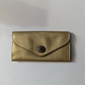 Puede incluir: Una cartera dorada con superficie texturizada y cierre de solapa. La cartera tiene un cierre de botón circular en el centro. La cartera rectangular parece estar hecha de cuero o material similar.