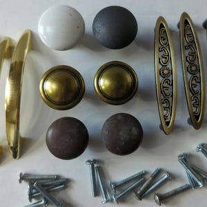 May include: Assortment of cabinet hardware including three gold-toned arched pulls, three gold-toned oval pulls with decorative detailing, two gold-toned round knobs, two brown round knobs, one white round knob, one gray round knob, and a collection of screws.