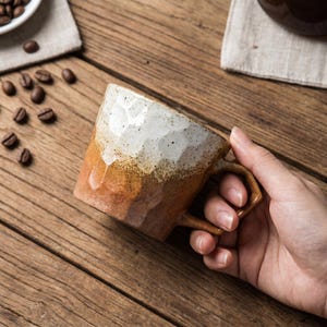 May include: A ceramic mug with a faceted, hammered texture featuring a two-tone glaze. The upper portion is a speckled off-white, transitioning into a warm, earthy brown at the base. The mug has a sturdy, angular handle and a cylindrical body.