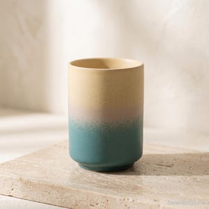 May include: A cylindrical ceramic cup featuring a smooth matte finish with a gradient colour transition. The top portion is a soft cream colour that blends into a muted pink midsection, which then fades into a deep teal base at the bottom.