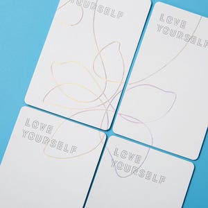 May include: Four white rectangular cards featuring the text LOVE YOURSELF in a thin, outlined font. Each card displays a delicate, continuous line drawing of a flower petal design in metallic gold, pink, and purple foil accents.