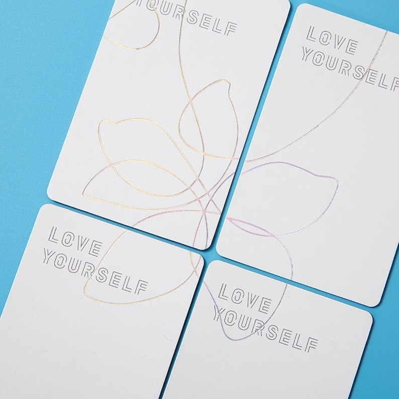 May include: Four white rectangular cards featuring the text LOVE YOURSELF in a thin, outlined font. Each card displays a delicate, continuous line drawing of a flower petal design in metallic gold, pink, and purple foil accents.