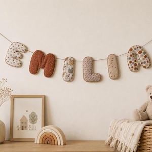 Op de afbeelding: Een stoffen letterslinger die de naam EMILIA spelt, bestaande uit gevulde letters in diverse patronen en effen kleuren, opgehangen aan een dun touwtje. De letters bevatten bloemen-, stippen- en effen roestkleurige stoffen ontwerpen.
