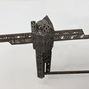 Op de afbeelding: Een donkergrijze, driedimensionale geometrische sculptuur met een ingewikkelde, roosterachtige structuur met herhalende zeshoekige en lineaire patronen. Het object heeft een centrale verticale kolom met horizontale uitbreidingen.