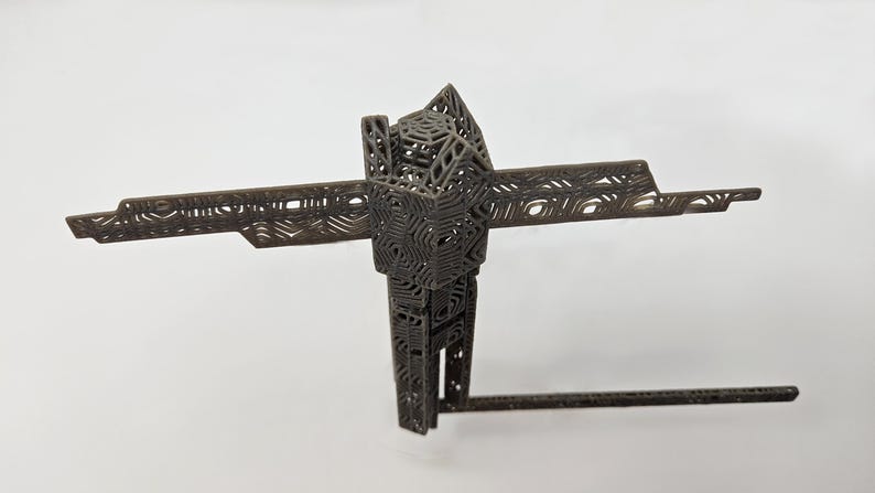 Op de afbeelding: Een donkergrijze, driedimensionale geometrische sculptuur met een ingewikkelde, roosterachtige structuur met herhalende zeshoekige en lineaire patronen. Het object heeft een centrale verticale kolom met horizontale uitbreidingen.