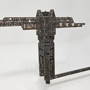 Op de afbeelding: Een donkerbruin, driedimensionaal object met een complexe, open roosterstructuur. Het heeft een centrale verticale kolom, een horizontale dwarsbalk aan de bovenkant en een enkele, lange, dunne arm die onder een hoek vanaf de basis uitsteekt.