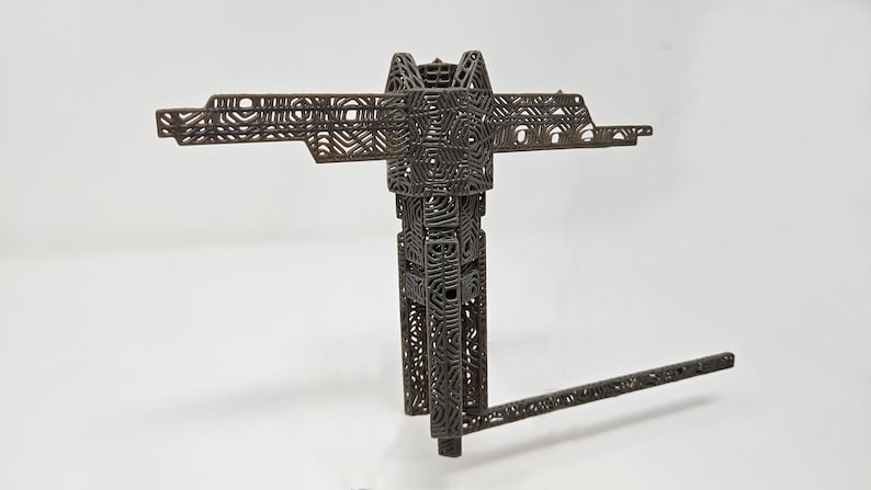 Op de afbeelding: Een donkerbruin, driedimensionaal object met een complexe, open roosterstructuur. Het heeft een centrale verticale kolom, een horizontale dwarsbalk aan de bovenkant en een enkele, lange, dunne arm die onder een hoek vanaf de basis uitsteekt.