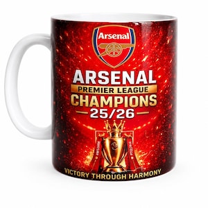 May include: A white ceramic mug featuring a red background with the Arsenal football club crest at the top. The text reads ARSENAL PREMIER LEAGUE CHAMPIONS 25/26 above a golden trophy illustration and the phrase VICTORY THROUGH HARMONY at the base.