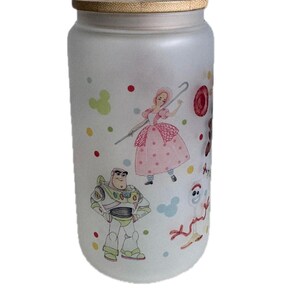 May include: A frosted glass tumbler with a bamboo lid featuring colorful illustrations of cartoon characters including a space ranger, a shepherdess with a staff, and a white spork character, accented by scattered red, yellow, and green polka dots.