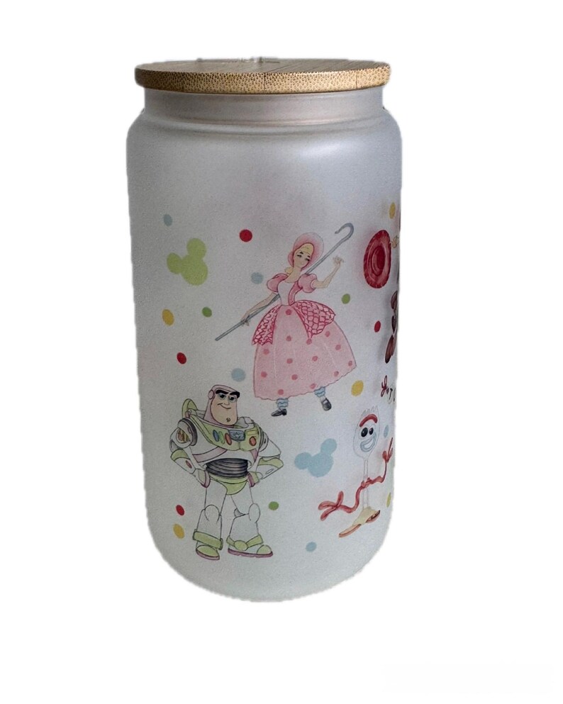 May include: A frosted glass tumbler with a bamboo lid featuring colorful illustrations of cartoon characters including a space ranger, a shepherdess with a staff, and a white spork character, accented by scattered red, yellow, and green polka dots.