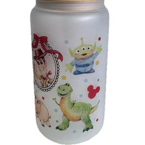 May include: A frosted glass tumbler with a bamboo lid featuring a colorful print of cartoon characters including a cowboy in a red-bowed frame, a green three-eyed alien, and a green dinosaur, accented by small multicolored dots and a red mouse icon.