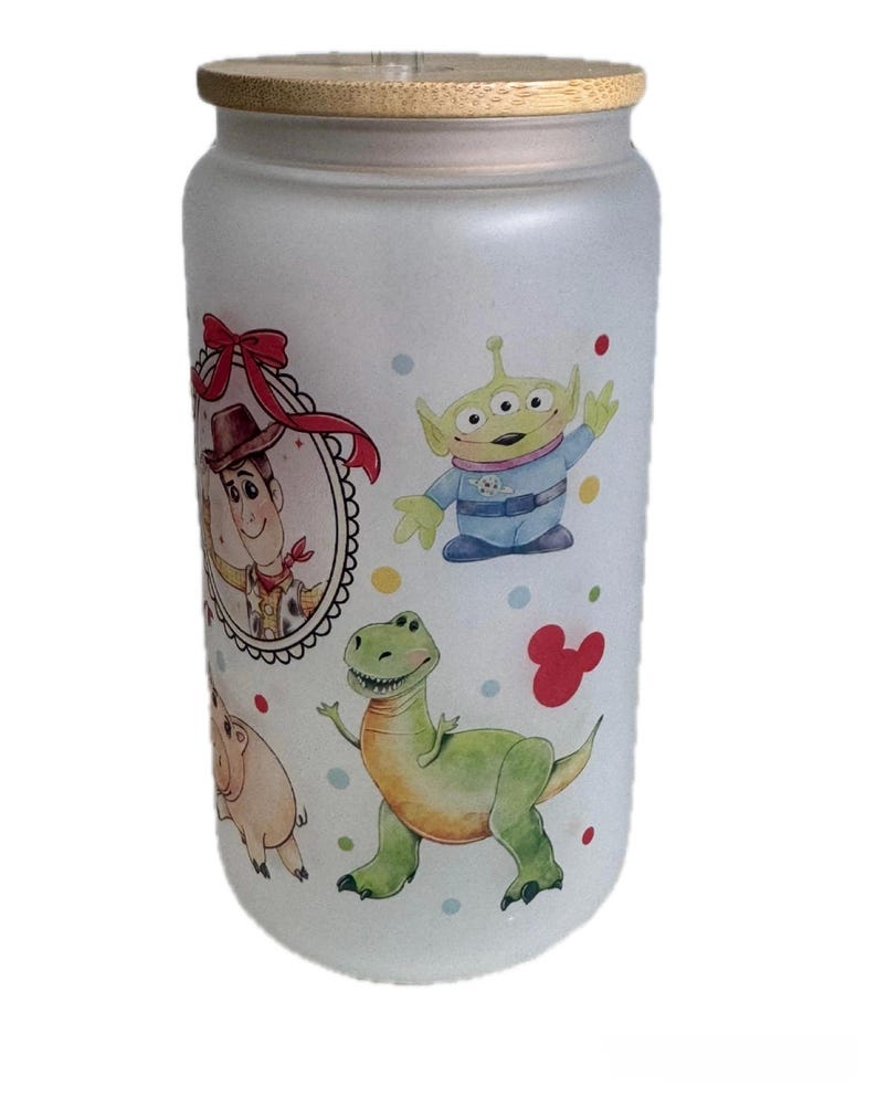 May include: A frosted glass tumbler with a bamboo lid featuring a colorful print of cartoon characters including a cowboy in a red-bowed frame, a green three-eyed alien, and a green dinosaur, accented by small multicolored dots and a red mouse icon.
