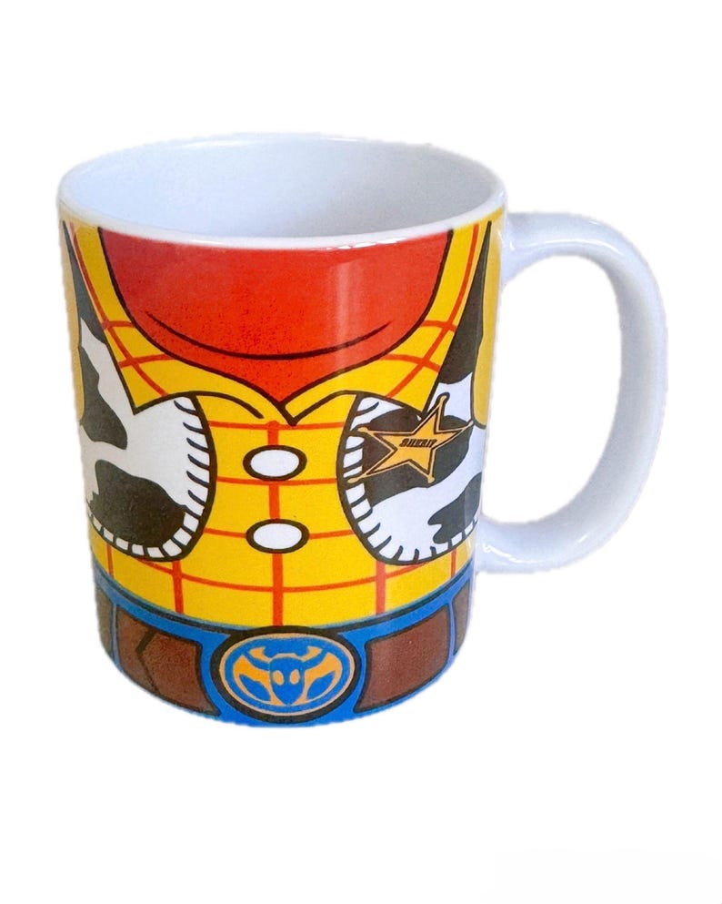 Puede incluir: Una taza de cer&aacute;mica blanca con un estampado gr&aacute;fico de una camisa de cuadros amarillos con un chaleco de manchas de vaca, un cintur&oacute;n marr&oacute;n con hebilla azul y una estrella de sheriff dorada. El dise&ntilde;o rodea el exterior de la taza con asa blanca.