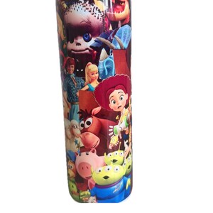 Puede incluir: Un vaso cil&iacute;ndrico de acero inoxidable con un estampado colorido que rodea todo el recipiente, mostrando varios personajes animados, incluyendo una vaquera de pelo rojo, un caballo marr&oacute;n, un cerdo rosa y alien&iacute;genas verdes de tres ojos.