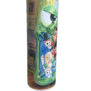 May include: A cylindrical metal tumbler featuring a colorful wrap-around illustration of various animated characters, including a large green one-eyed creature, a green dinosaur, and several white spork-like figures with googly eyes.