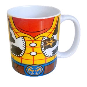 Puede incluir: Una taza de cer&aacute;mica blanca con un estampado gr&aacute;fico de una camisa de cuadros amarillos con un chaleco de manchas de vaca, un cintur&oacute;n marr&oacute;n con hebilla azul y una estrella de sheriff dorada. El dise&ntilde;o rodea el exterior de la taza con asa blanca.