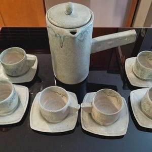 May include: A ceramic tea set featuring a tall cylindrical teapot with a side handle and lid, accompanied by six matching cups on square saucers. The set has a mottled, pale green and off-white glaze with visible drip patterns on the teapot.