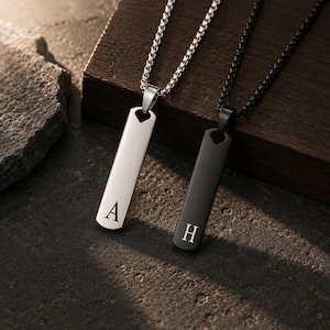 May include: Two rectangular pendant necklaces with box chains. One pendant is silver-toned with the letter A engraved at the bottom, and the other is black with the letter H engraved at the bottom. Both pendants feature a small heart-shaped cutout.