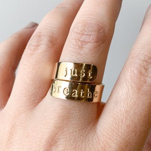 May include: Two gold rings with the words "just breathe" engraved on them. The rings are worn on a finger.
