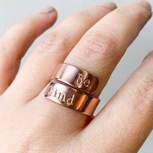 May include: A rose gold-colored ring with the words "be kind" engraved on it. The ring is worn on a person's finger.