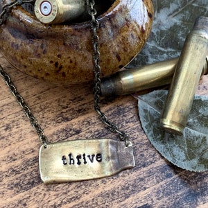 May include: A brass necklace with a pendant that reads "thrive". The pendant is made from a repurposed bullet casing. The necklace is displayed on a wooden surface with a ceramic bowl filled with bullet casings.