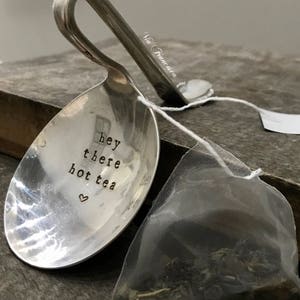 May include: A silver spoon with a handle bent into a loop, engraved with the text "hey there hot tea" and a heart. A tea bag is in the background.